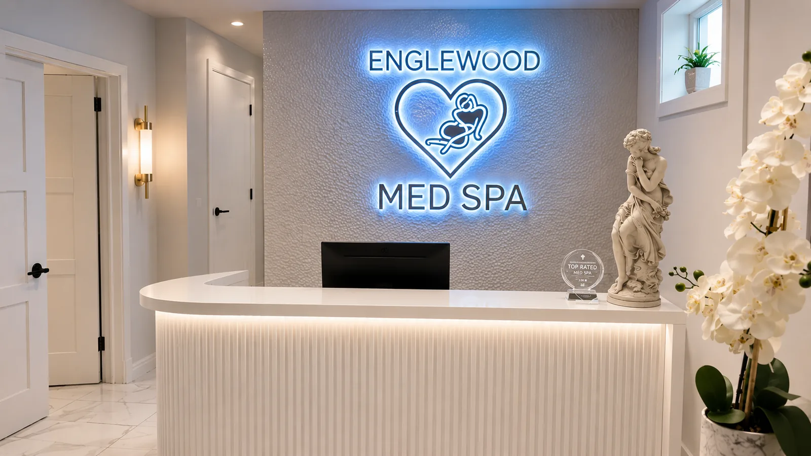 Reception desk at Englewood Cosmetic Medical Spa with a backlit ENGLEWOOD MED SPA sign, white marble floors and orchids
